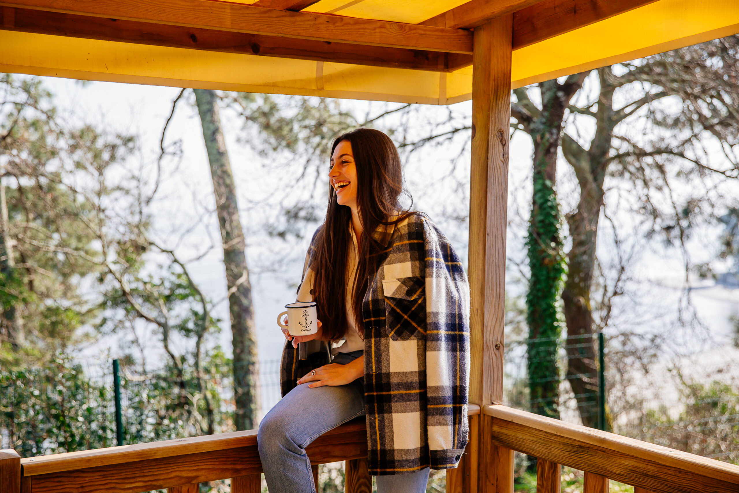 Une femme boit un thé, assise sur la rambarde d'un mobil-home au camping Les Pastourelles, avec le Bassin en arrière-plan.