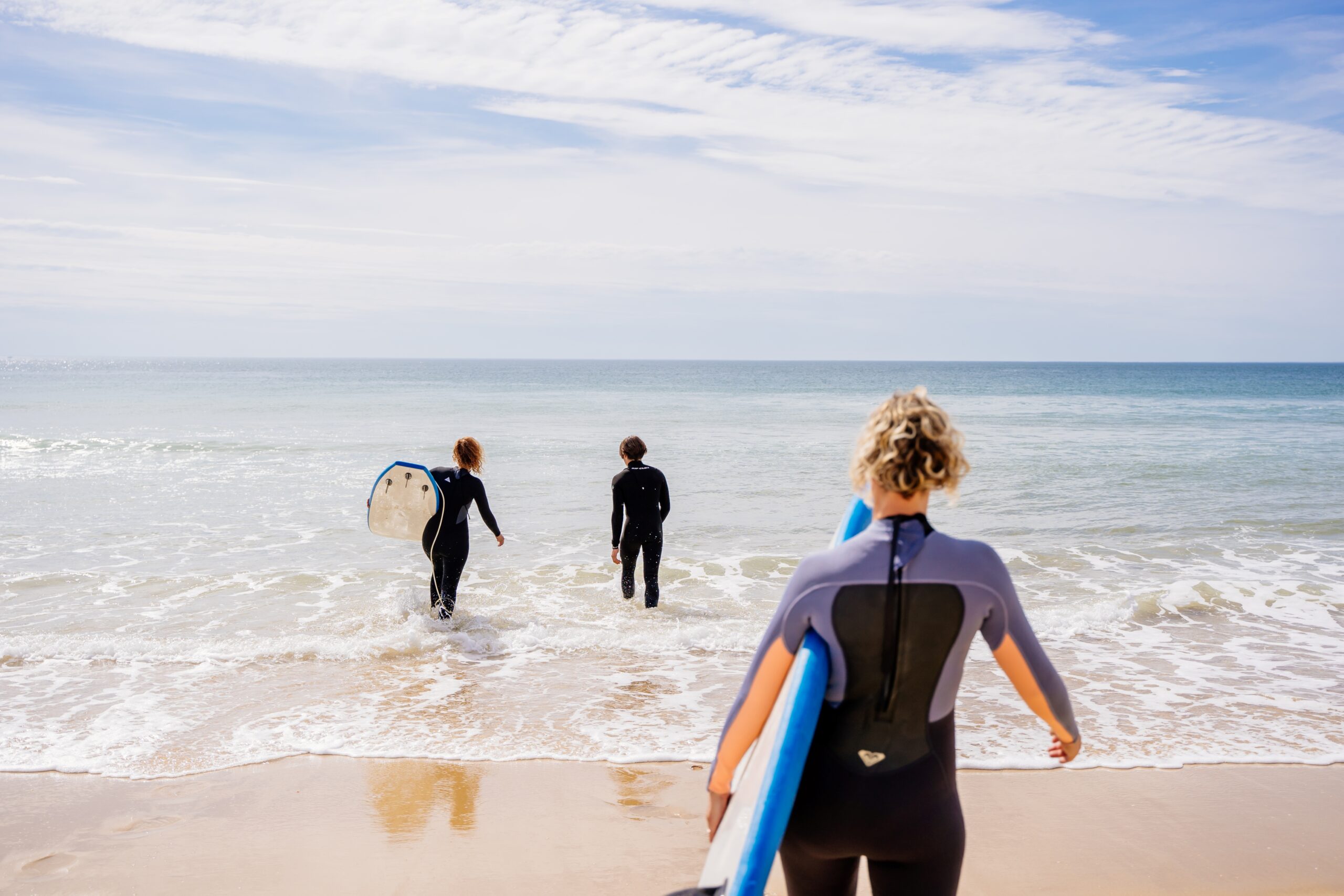 Trois amis novices en surf entrent dans l'eau, planche à la main, prêts à affronter les vagues.