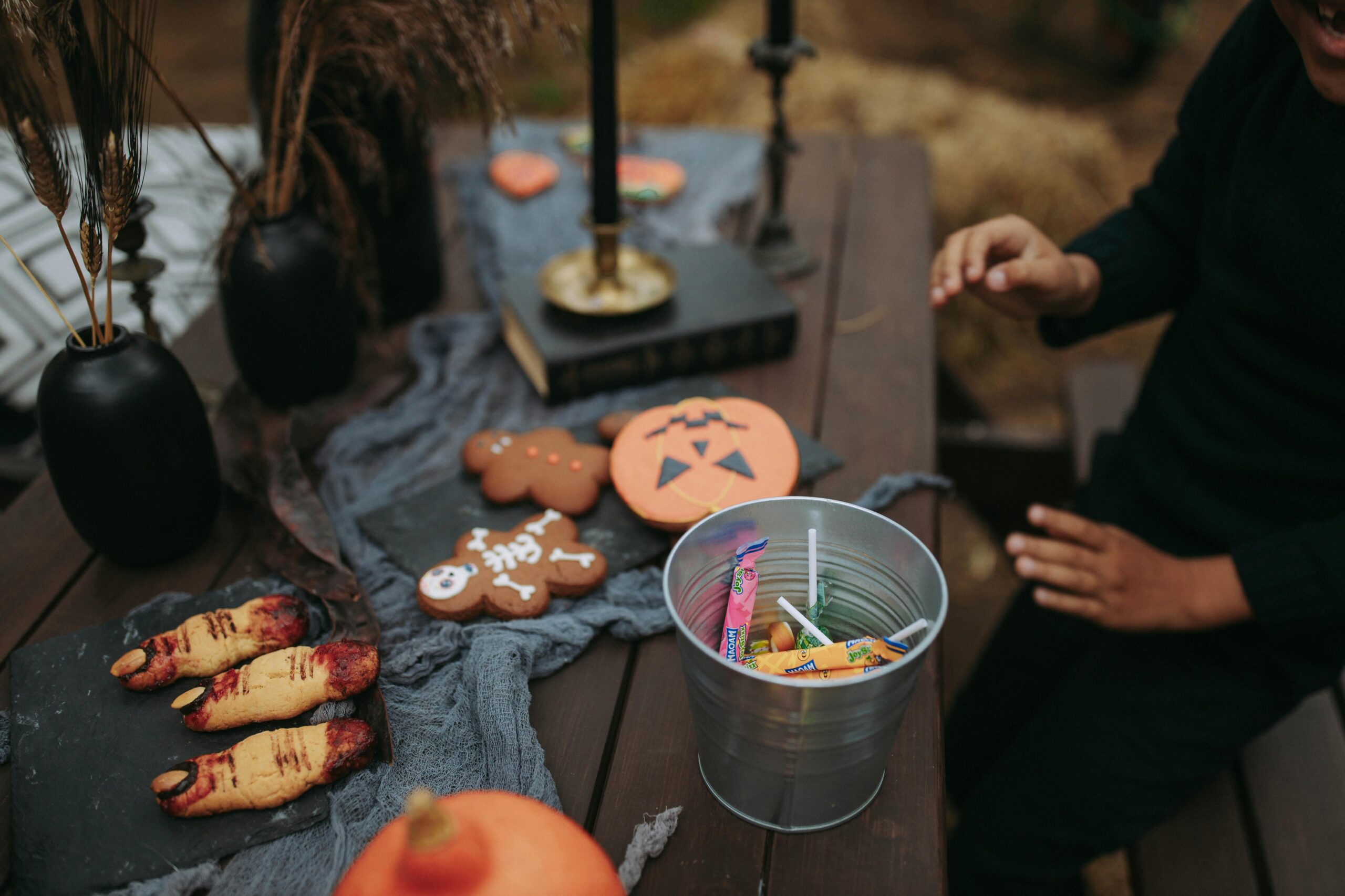 Table de fête Halloween décorée avec des biscuits en forme de citrouille, de squelette et de doigts ensanglantés, un seau en métal rempli de bonbons, et des chandeliers noirs, créant une ambiance festive et effrayante pour Halloween.