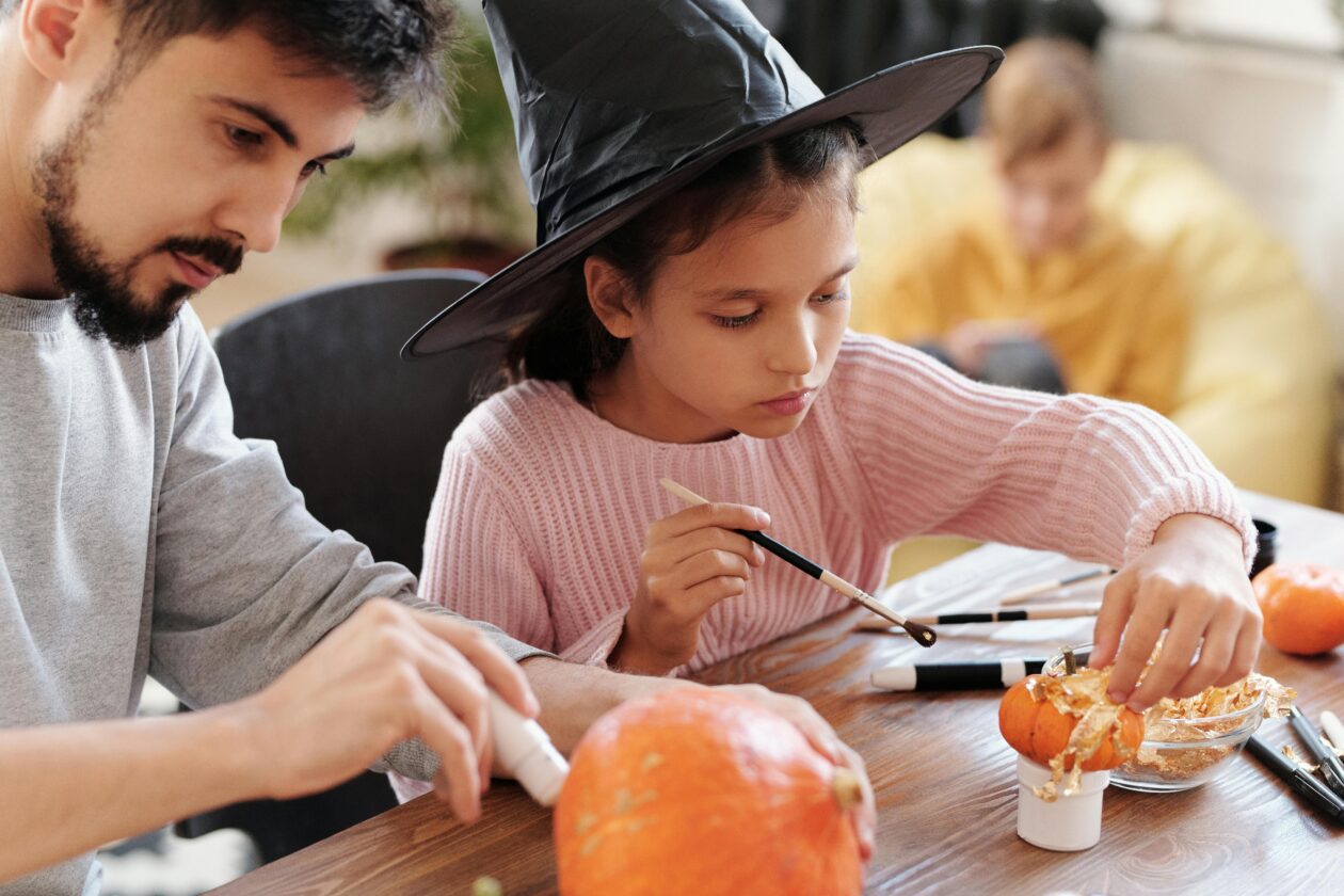 Un homme et une jeune fille décorent des citrouilles pour Halloween, la fille portant un chapeau de sorcière et utilisant un pinceau, entourés de paillettes et d'outils d'artisanat sur une table.