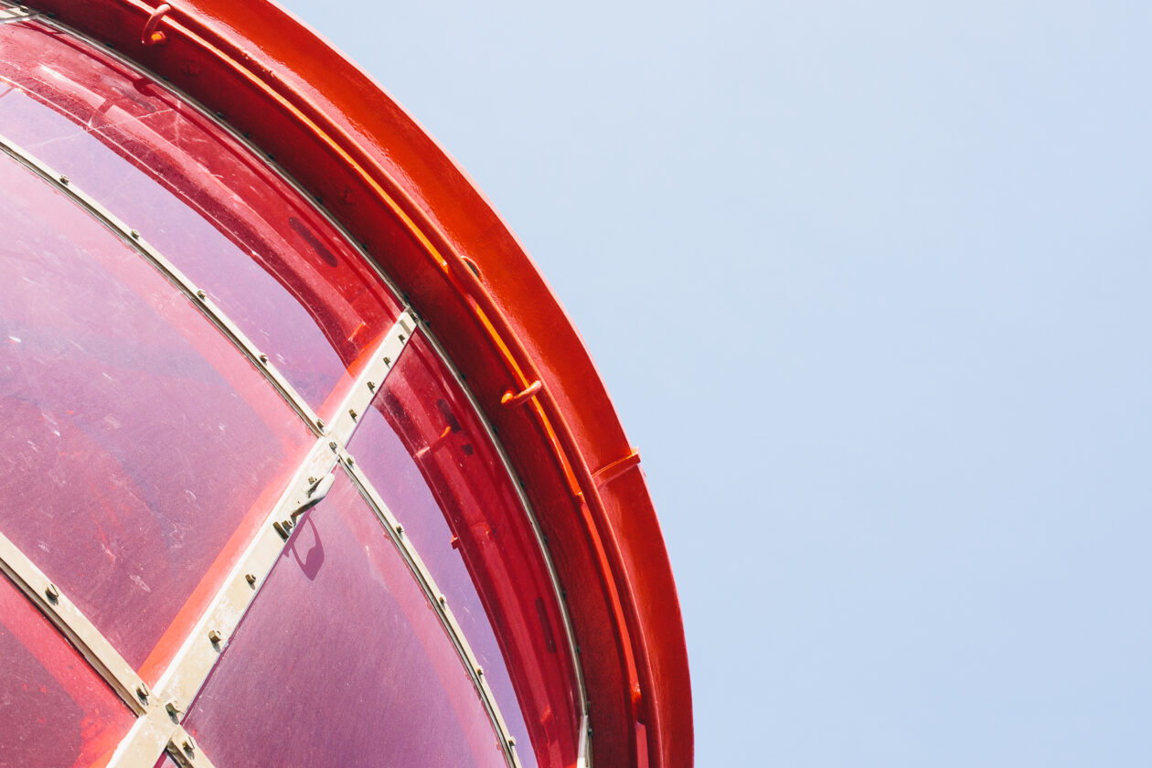  Gros plan en contre‑plongée sur la lanterne rouge d’un phare : la structure cylindrique, soutenue par des barres métalliques et des rivets, se détache sur un ciel bleu limpide. 